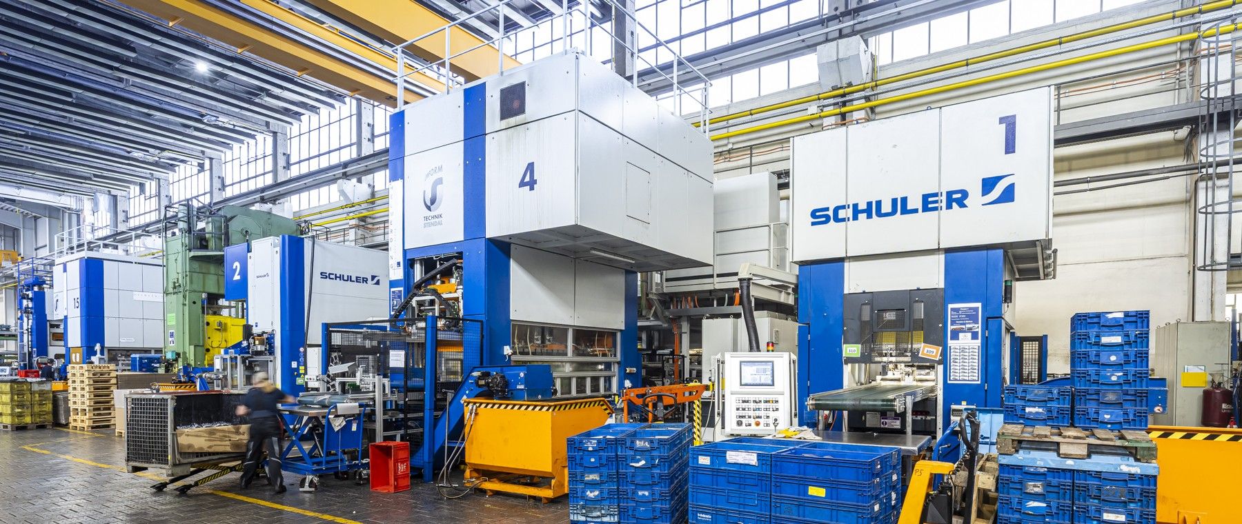 Innenraum einer Industrieanlage mit großen Schuler-Maschinen, blauen Lagerbehältern und einem Arbeiter, der Materialien auf dem Boden transportiert.