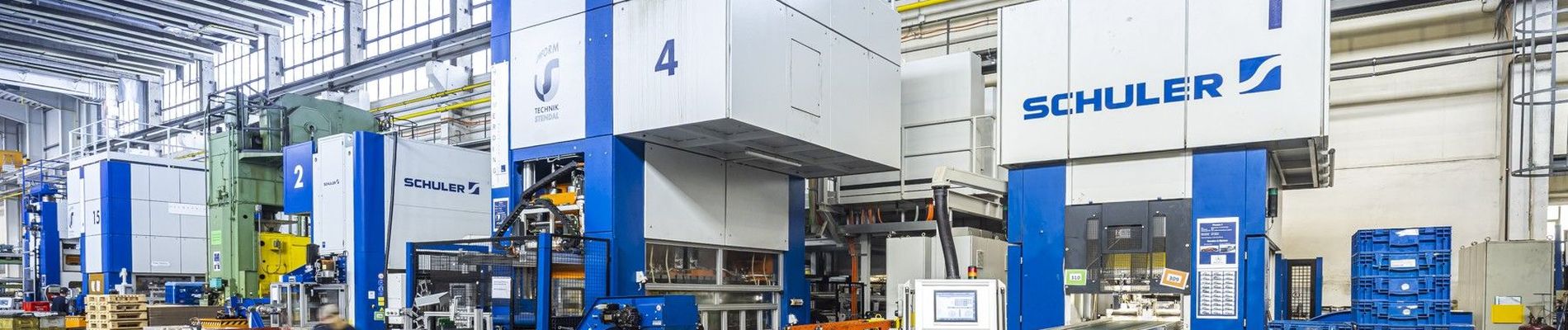 Innenraum einer Industrieanlage mit großen Schuler-Maschinen, blauen Lagerbehältern und einem Arbeiter, der Materialien auf dem Boden transportiert.